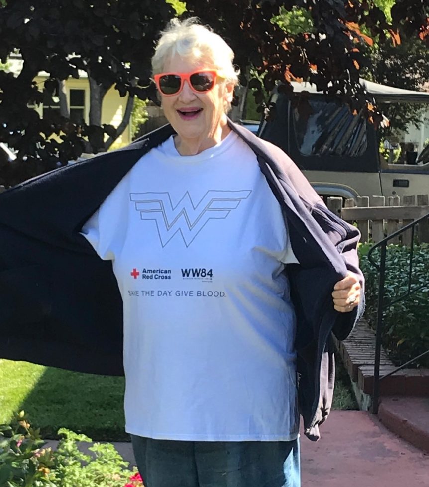 smiling elderly woman wearing sunglasses opening her coat to reveal t-shirt with a wonder woman logo and text that says "save the day, give blood"
